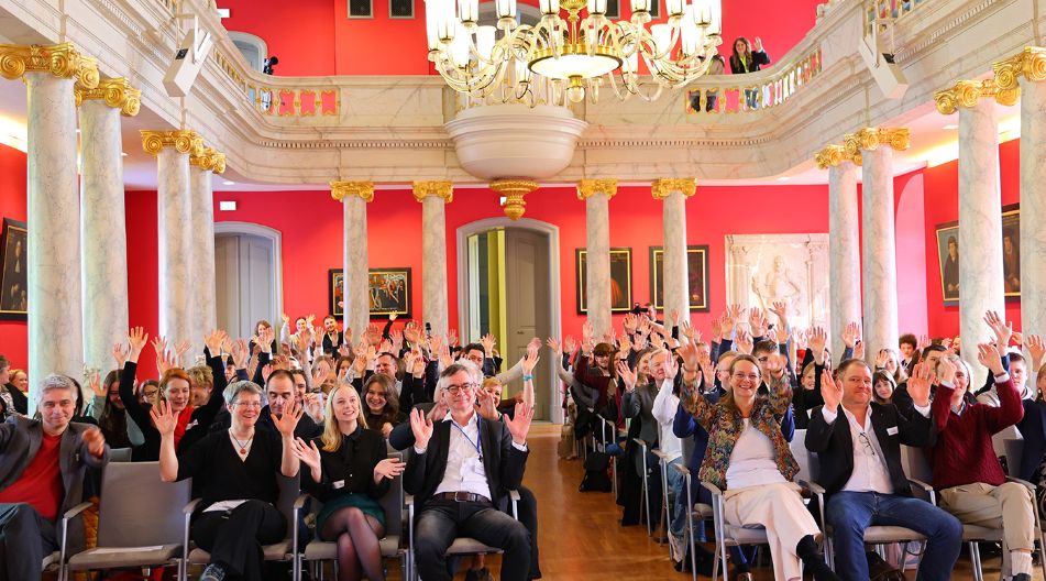 Ein festlicher Saal mit roten Wänden, goldverzierten Säulen und einem großen Kronleuchter. Eine große Gruppe von Menschen sitzt auf Stuhlreihen, lächelt und hebt die Hände in die Luft. Einige Personen stehen auf der Empore und winken.
