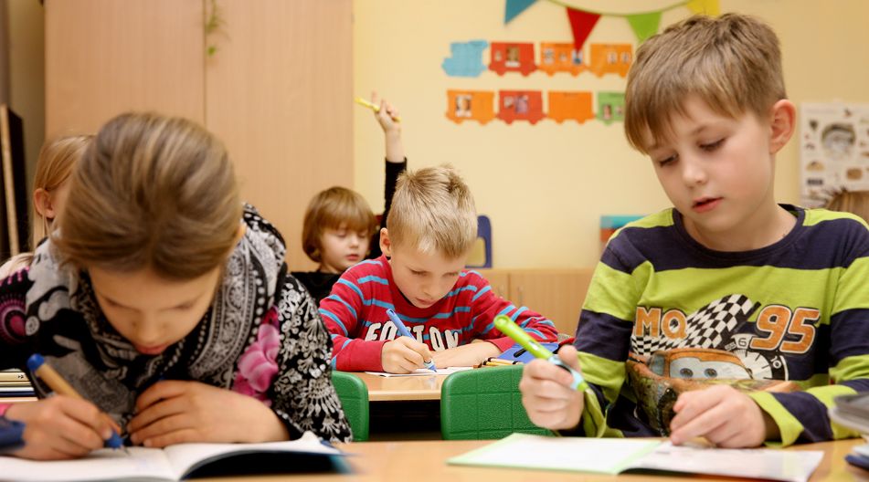 Mehrere Grundschulkinder sitzen in einem bunt dekorierten Klassenzimmer an ihren Tischen und schreiben konzentriert in ihre Hefte. Ein Kind im Hintergrund hebt die Hand. Mehrere Grundschulkinder sitzen in einem bunt dekorierten Klassenzimmer an ihren Tischen und schreiben konzentriert in ihre Hefte. Ein Kind im Hintergrund hebt die Hand.